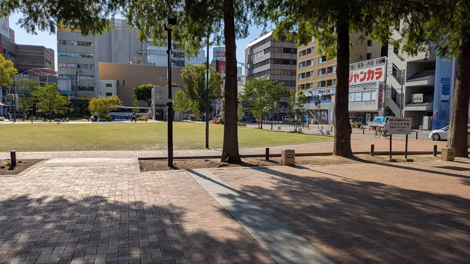 阪神尼崎駅前・中央公園の芝生広場と木陰の石畳。周囲に駅前のビルが見える