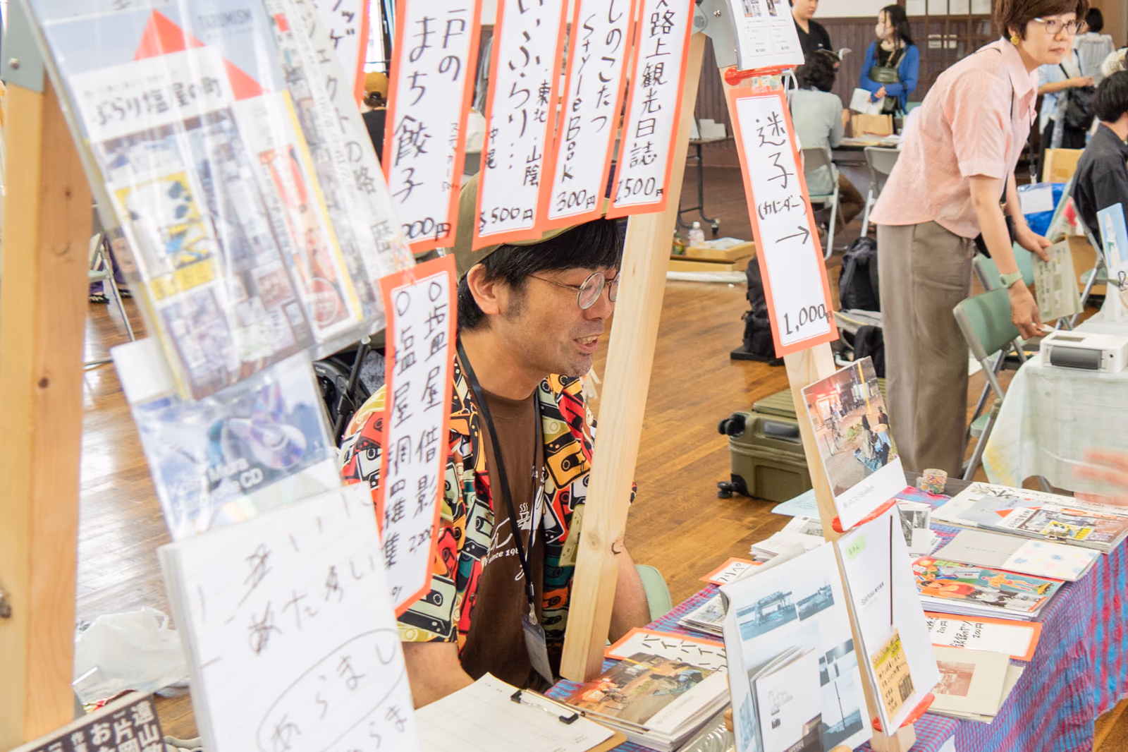 一箱古本市のブースで、手書きの値札を吊るした冊子に囲まれて座るリトルレンズ文芸舎