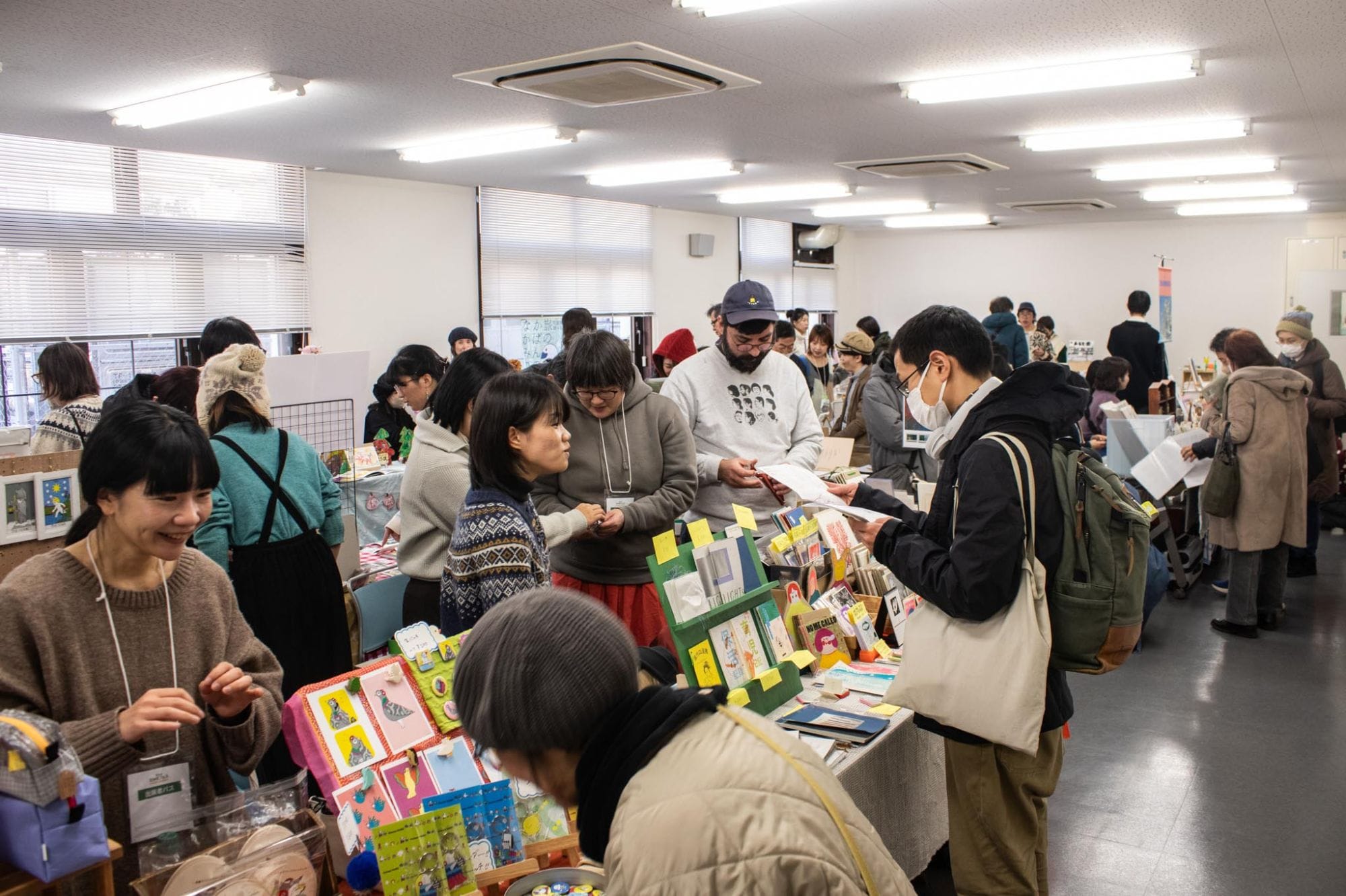 まちのZINEフェスの会場。長机に並んだZINEや雑貨を手に取る来場者と、笑顔で迎える出展者たち